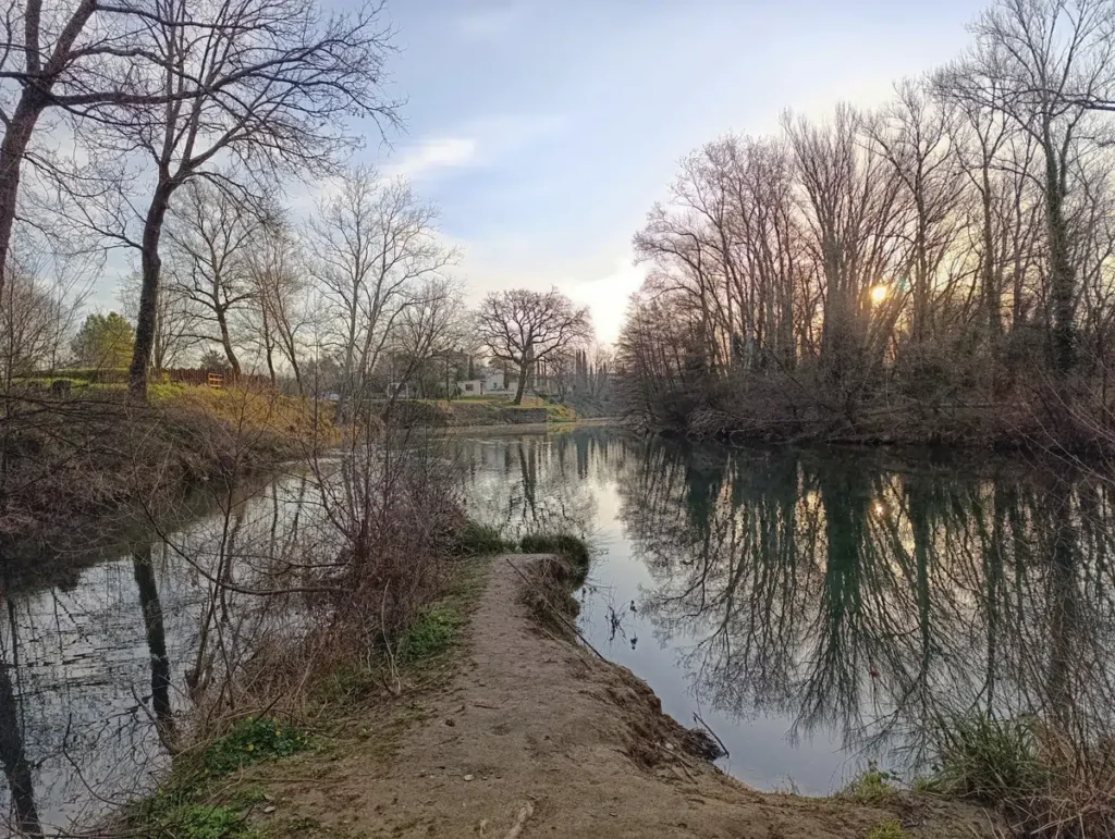 Photo d'un paysage avec un petit fleuve à l'eau calme bordée d'arbres principalement dépourvus de feuilles. Un sentier étroit avance vers l'eau au premier plan. La rivière reflète les arbres et le ciel. À l'arrière-plan, une maison claire avec un toit sombre est visible derrière une petite butte herbeuse, partiellement cachée par des arbres. Le soleil est bas et partiellement masqué par les branches, projetant une lumière diffuse.