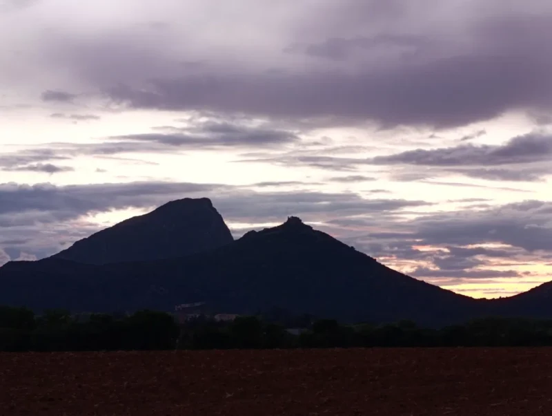 Un paysage de fin de journée, avec un premier plan constitué d’un champ de terre nue et sombre. Derrière ce champ, une bande de végétation arbustive sépare le sol cultivé des reliefs montagneux.
Deux sommets se détachent sur l’horizon : l’un, plus massif, à gauche, le Pic Saint Loup, et l’autre, plus effilé, à droite, avec un petit château en ruine à peine visible au sommet du second. Le ciel est largement couvert de nuages.