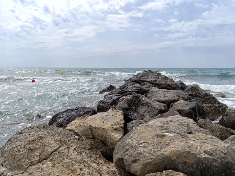 Une digue de rochers s'avance dans une mer légèrement agitée sous un ciel voilé de nuages. Sur la gauche de la photo, deux bouées jaunes et une bouée orange indiquent un chenal.