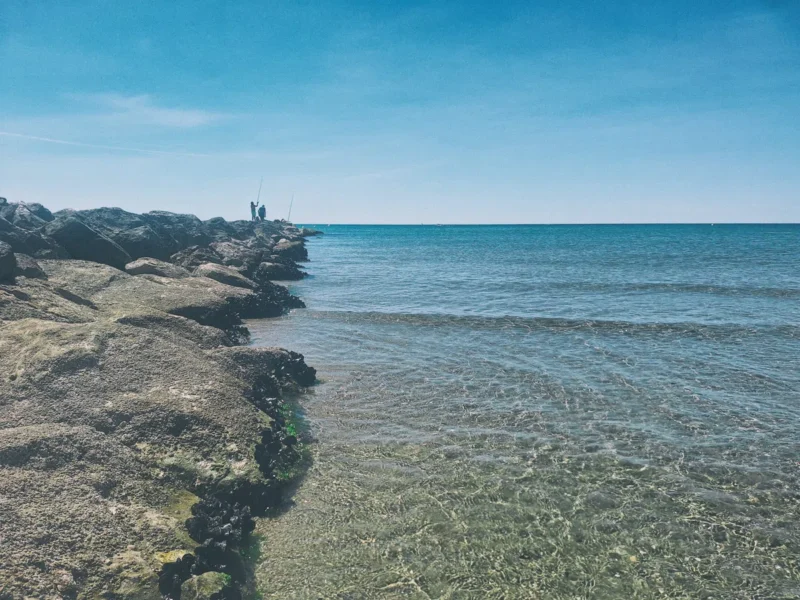 Mer calme sous un ciel bleu clair. Au premier plan, sur la gauche, des rochers forment une jetée s’avançant dans l’eau. Deux personnes debout sur la jetée tiennent des cannes à pêche. L’eau est claire et peu profonde près du rivage.