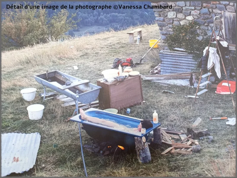 Photo. Sur un terrain en pente herbeuse près d’un bâtiment en pierre. Une femme est allongée dans une baignoire bleue, remplie d’eau, posée sur deux troncs de bois, avec un feu allumé en dessous. Autour de la baignoire, plusieurs objets sont disposés : planches de bois, bouteilles en plastique, bassines, ustensiles de cuisine, une bassine métallique sur pieds, un meuble en bois servant de plan de travail, des seaux, des vêtements suspendus et des éléments de bricolage. À l’arrière-plan, un paysage vallonné et boisé.