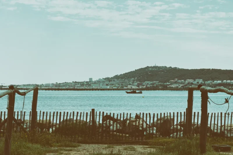 Vue sur un étang avec une ville (Sète) en partie sur une colline boisée. Au centre de la photo, un bateau noir est visible sur l’eau. À l’avant-plan, une clôture en bois brut encadre la scène.