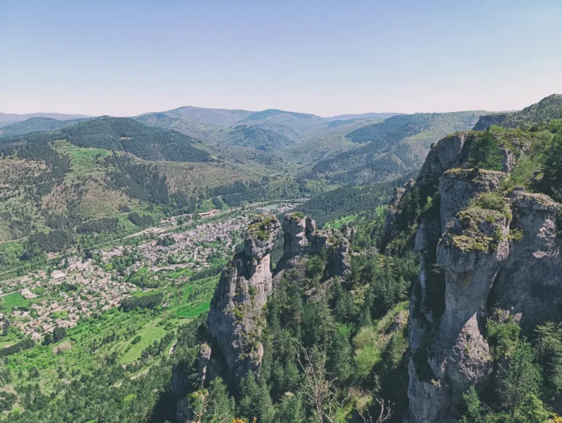 Un paysage montagneux avec une vue panoramique sur une vallée. À droite, des falaises rocheuses escarpées dominent le paysage, couvertes de végétation. En contrebas, une petite ville ou un village est niché au milieu de la verdure, avec des maisons dispersées. Au loin, des collines et des montagnes boisées s'étendent sous un ciel clair et bleu.