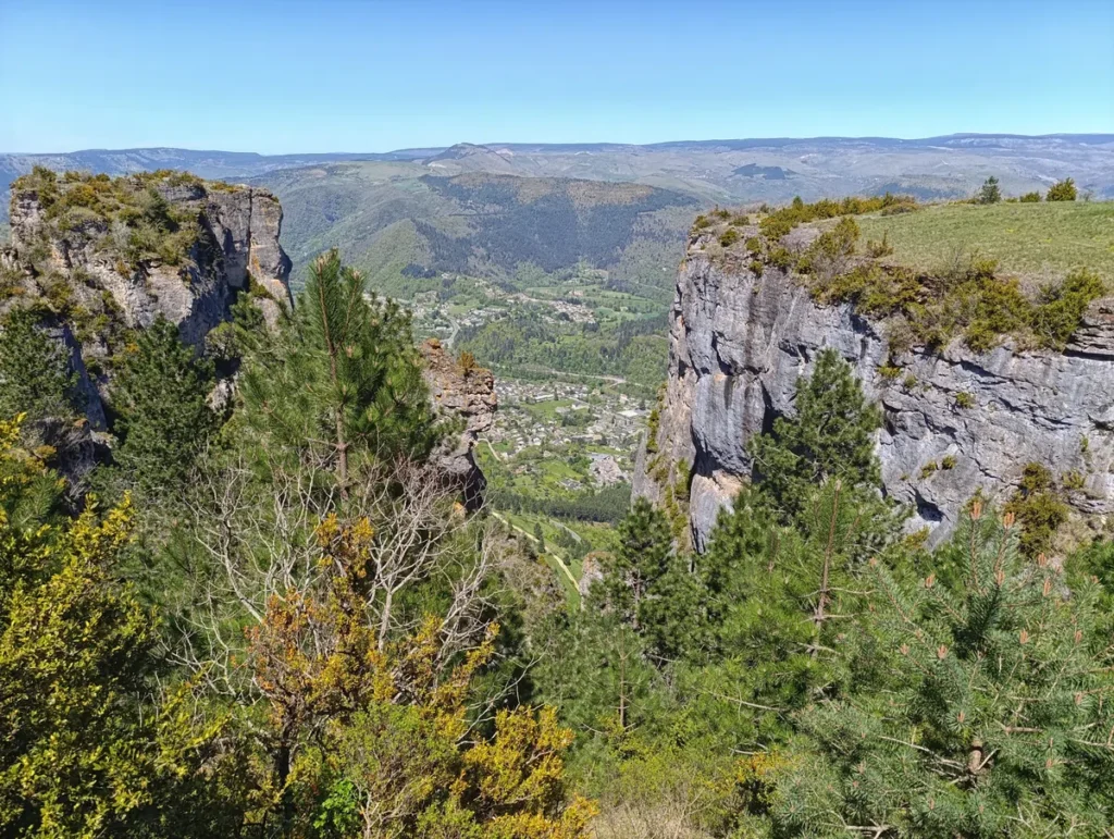 Un paysage montagneux avec des falaises rocheuses au premier plan, entourées de végétation, notamment des arbres et des arbustes. En contrebas, une vallée s'étend avec un village niché au milieu de la verdure. À l'arrière-plan, des collines et des montagnes boisées se succèdent sous un ciel bleu clair.