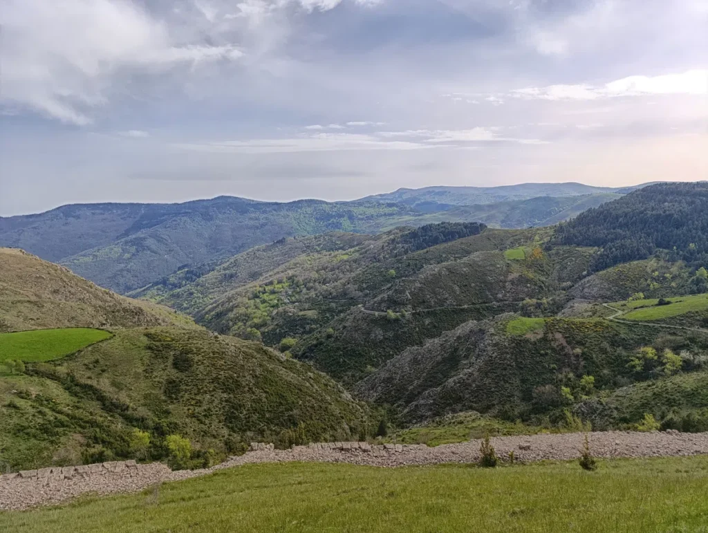 Un paysage de montagnes et de vallées sous un ciel partiellement nuageux. Au premier plan, il y a une zone herbeuse avec un muret de pierres sèches. Les collines et les montagnes à l'arrière-plan sont couvertes de végétation, avec des zones boisées et des prairies. L'horizon montre des montagnes plus éloignées sous un ciel lumineux.