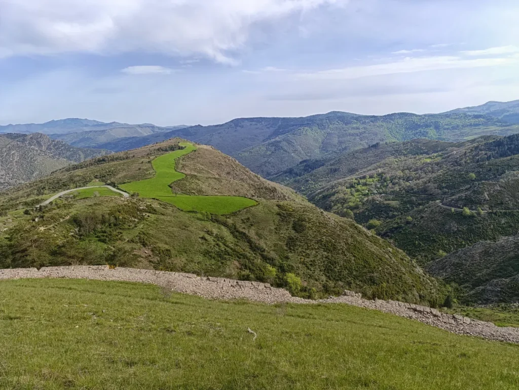 Un paysage de collines et de montagnes sous un ciel partiellement nuageux. Au premier plan, il y a une zone herbeuse avec un muret de pierres sèches. Les collines sont couvertes de végétation, avec des prairies vertes et des zones boisées. À l'arrière-plan, des montagnes s'étendent à l'horizon, offrant une vue panoramique sur la nature.