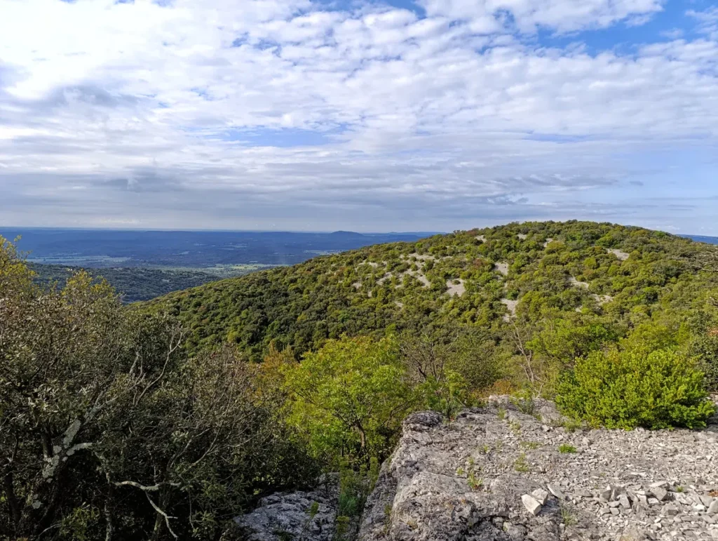 Paysage de montagne avec une colline boisée au centre, vue depuis un promontoire rocheux au premier plan, sous un ciel partiellement nuageux laissant entrevoir des zones de ciel bleu.