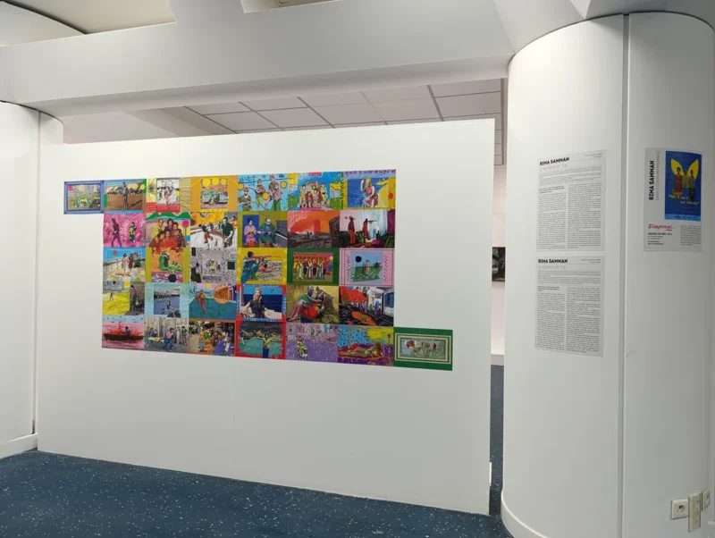 Photo d'une salle d’exposition. Une cloison blanche est utilisée pour présenter une série de compositions visuelles disposées en grille. Des photos coloriées, comportant des couleurs vives et représentant des scènes variées, souvent humaines ou urbaines. À droite, sur un pilier cylindrique, trois cartels.