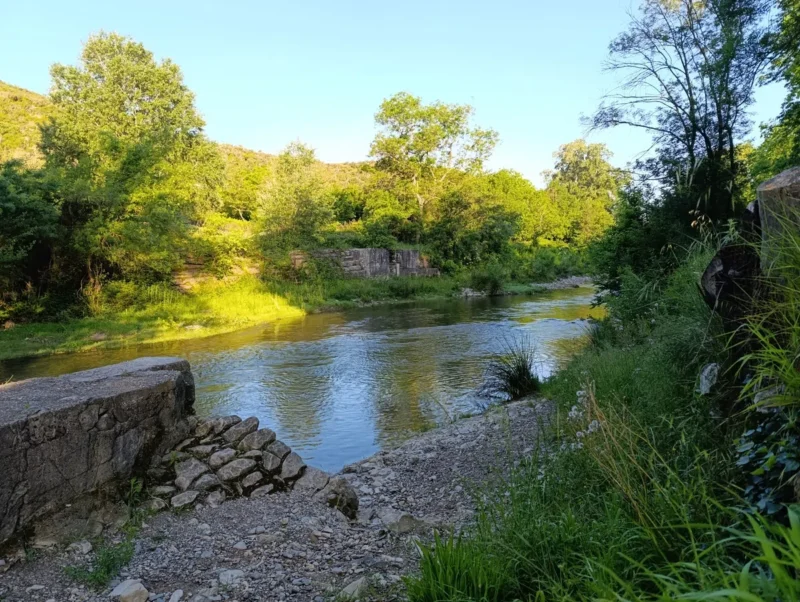 Un petit fleuve au petit matin, avec quelques zones plus écrasées par le soleil, d'autres encore à l'ombre. Sur les rives, de la végétation et d'anciens vestiges de murs et d'escaliers qui permettaient d'accéder au cours d'eau. Le ciel est bleu, sans nuage.