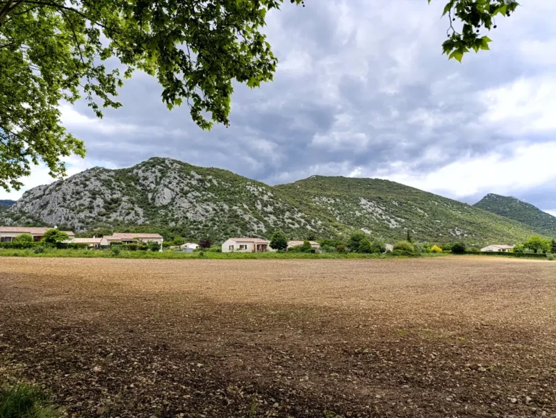 À l’avant-plan, un champ de terre nue fraîchement labouré. En arrière-plan, une rangée de maisons alignées au pied d’un massif calcaire couvert de végétation. Au-dessus, le ciel est couvert de nuages. La partie supérieure de l’image est partiellement encadrée par les branches feuillues d’un arbre.