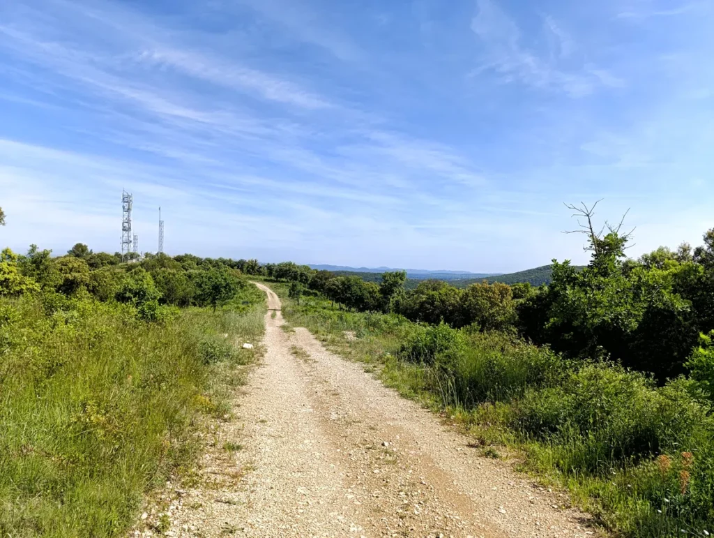 Un chemin sur un petit massif à la végétation basse. Sur la gauche, on perçoit des émetteurs (téléphones et télé). Au-dessus, le ciel est voilé de quelques tracés nuageux.