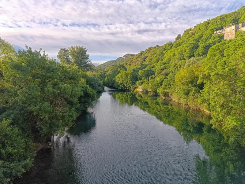 Vue d’un petit fleuve bordé de collines boisées. À droite, des maisons en pierre sont visibles sur les hauteurs. Le ciel est partiellement couvert de nuages.