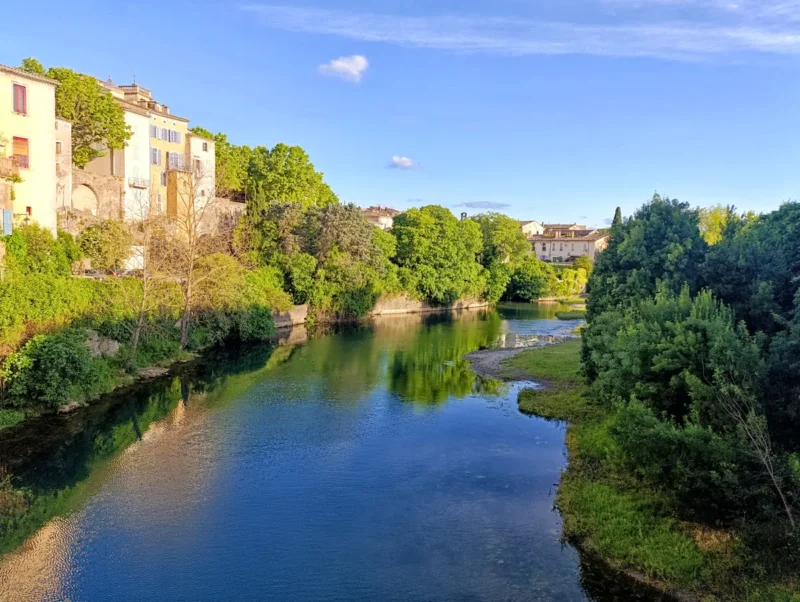 Vue d’un petit fleuve traversant un village. À gauche, des bâtiments aux façades colorées longent l’eau. À droite, des arbres et des berges herbeuses bordent le cours d'eau. Le ciel est dégagé avec quelques nuages.