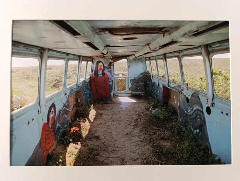 Photo d'une photographie d'artiste bordée de blanc. Intérieur d’un véhicule (peut-être un bus) abandonné au milieu de la nature avec à l'intérieur des peintures murales représentant des visages de femmes. Le sol du véhicule est recouvert de sable et de végétation. Des plantes sont visibles à l’extérieur à travers les fenêtres.