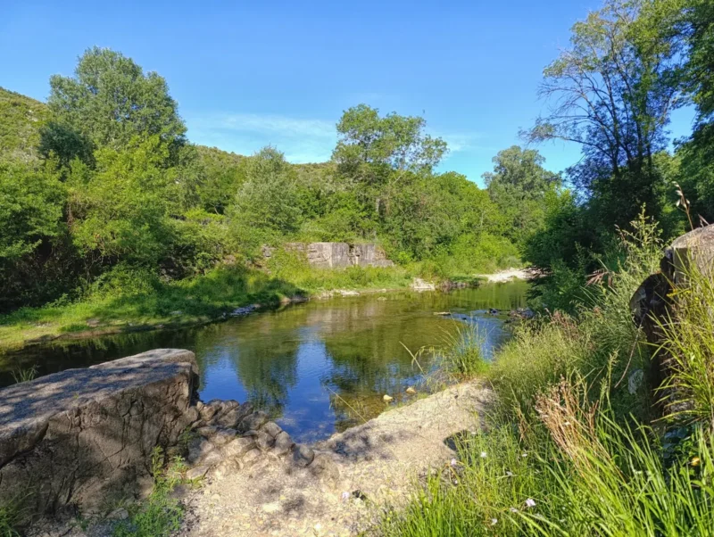 Sous les bords d'un petit fleuve, sous un ciel bleu. Au premier plan, sur la gauche, les vestiges d'un mur, une ancienne construction. Idem sur la rive opposée. De l'herbe bien verte sur la droite de la photo. Des arbres et de la végétation aux airs printaniers sur les rives du paisible cours d'eau.