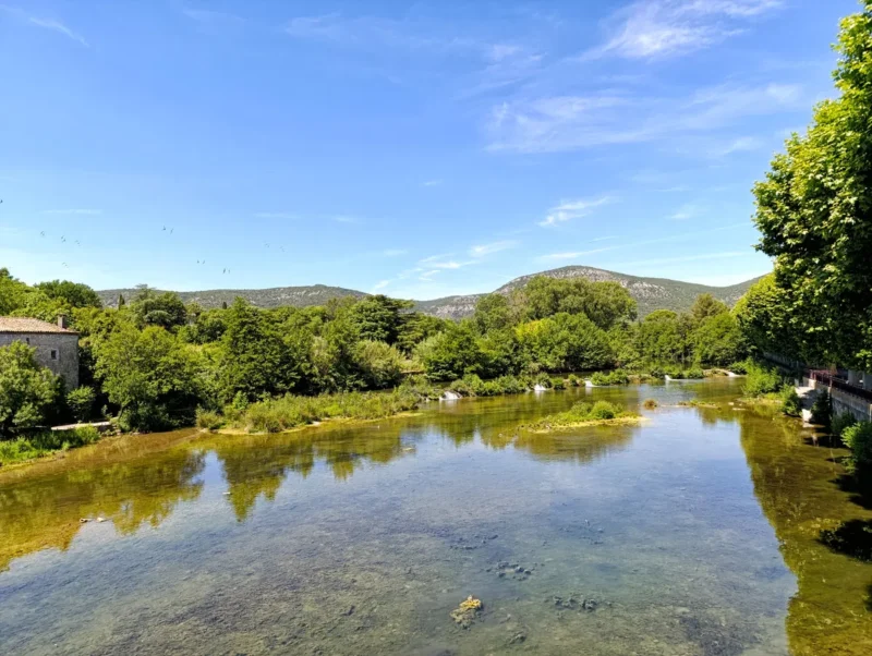 Photo d’un cours d'eau peu profond bordé de végétation, avec des arbres sur les deux rives. À gauche, une maison en pierre partiellement visible. En arrière-plan, des collines boisées sous un ciel bleu clair avec quelques nuages fins. Sur la droite, une rangée de platanes aux feuillages denses. On perçoit quelques traces floues de vols d'hirondelles dans le ciel, sur la gauche de l'image.