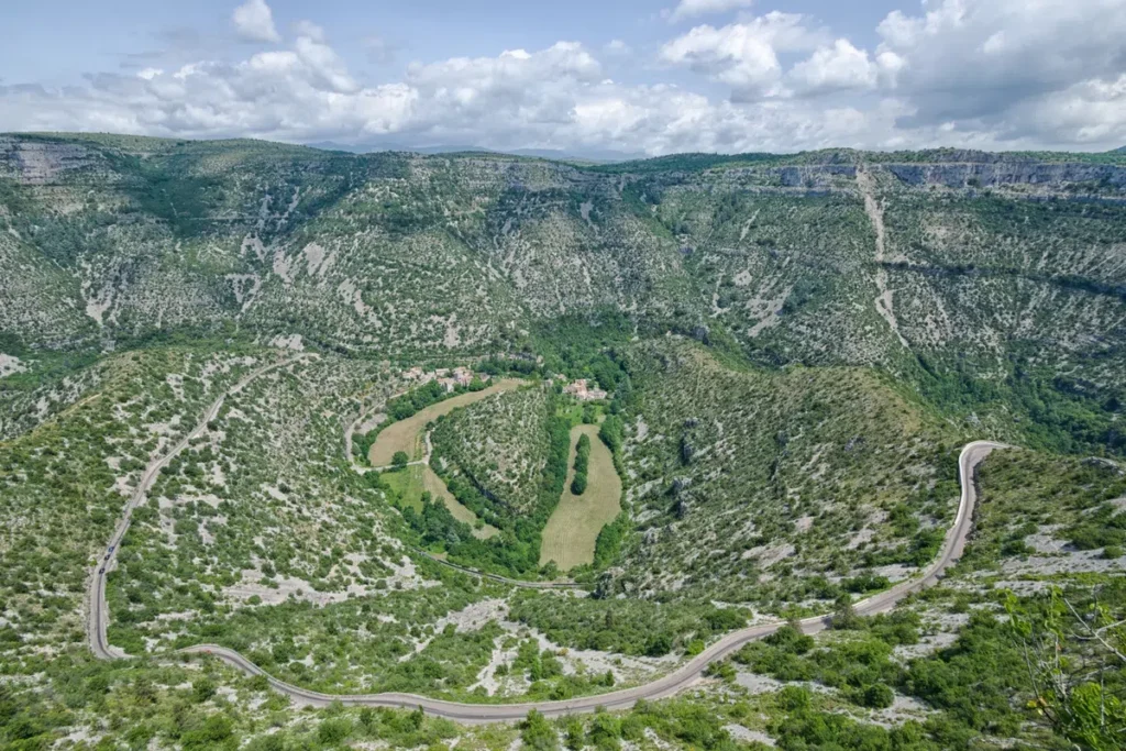 Vue aérienne du Cirque de Navacelles, une vallée encaissée entourée de montagnes recouvertes de végétation clairsemée. Une route sinueuse descend vers un hameau niché au fond de la vallée, bordé de champs en forme de méandre.