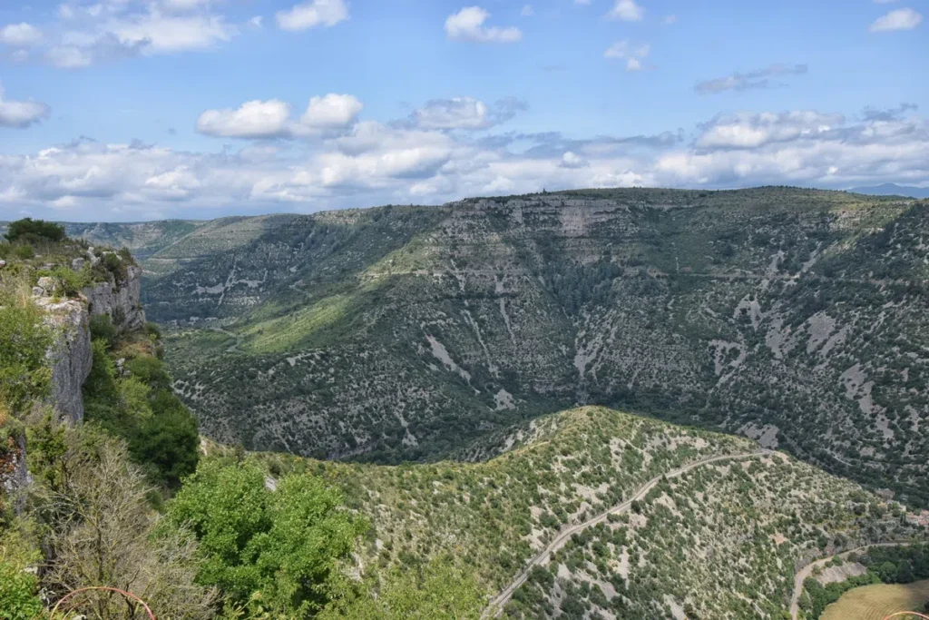 Vue panoramique du Cirque de Navacelles. Parois rocheuses et végétation éparse. Une route sinueuse longe la crête au premier plan, avec un ciel partiellement nuageux en arrière-plan.
