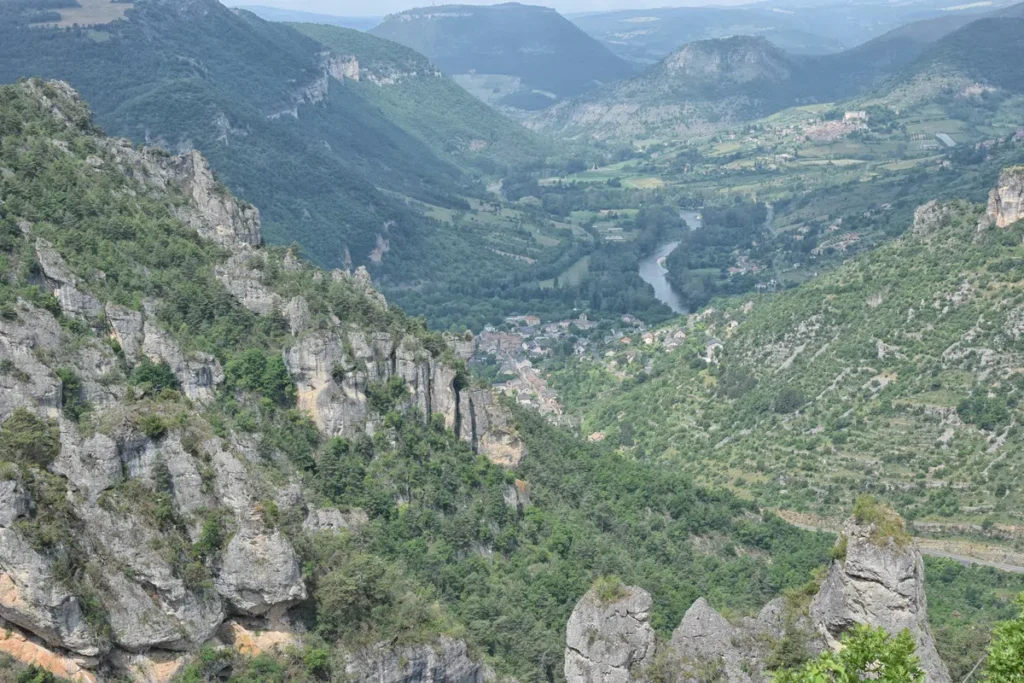 Photo d'un paysage de montagnes et de vallées. Au premier plan, on voit des falaises rocheuses couvertes de végétation verte. En descendant, la vue s'ouvre sur une vallée verdoyante avec une rivière sinueuse qui serpente à travers des champs et des zones boisées. Au loin, on aperçoit un petit village niché parmi les arbres, avec des maisons regroupées. Les montagnes environnantes sont couvertes de forêts denses et s'étendent jusqu'à l'horizon sous un ciel légèrement brumeux.