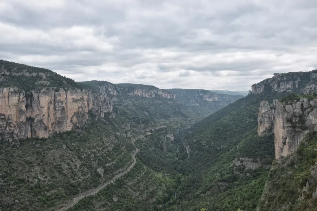 Photo d'un paysage de montagnes et de vallées. On y voit des falaises rocheuses imposantes de chaque côté, couvertes de végétation verte. Une route sinueuse serpente à travers la vallée, disparaissant dans le lointain. Le ciel est couvert de nuages gris. La vallée est dense avec des arbres et des buissons, et les montagnes s'étendent jusqu'à l'horizon.