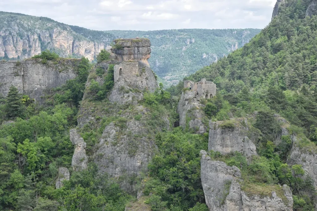 Photo d'un paysage de montagnes avec des ruines de bâtiments anciens. Au centre, on voit les vestiges d'une structure en pierre, un ermitage, perchée sur un éperon rocheux. Les ruines sont entourées de végétation dense et de falaises abruptes. Le ciel est partiellement couvert de nuages, et les montagnes environnantes sont couvertes de forêts vertes.