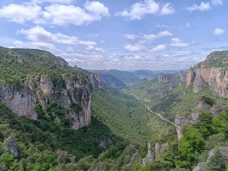 Photo d'un paysage de montagnes et de vallées. On y voit des falaises rocheuses couvertes de végétation verte. Une route sinueuse serpente à travers la vallée, disparaissant dans le lointain. Le ciel est partiellement couvert de nuages blancs, laissant entrevoir des zones de ciel bleu. Les montagnes environnantes sont couvertes de forêts denses et s'étendent jusqu'à l'horizon.