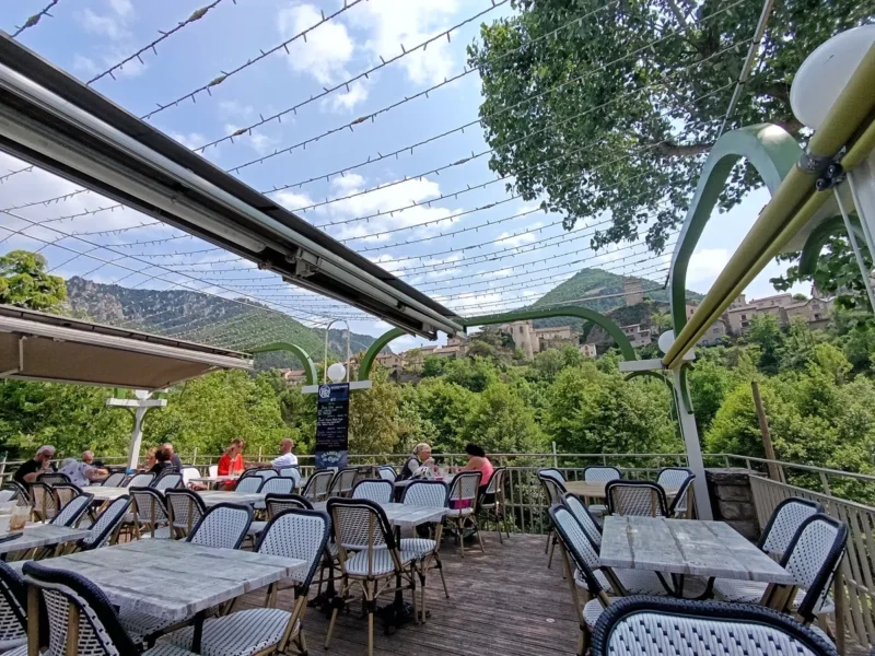 Vue d'une terrasse de café avec quelques personnes attablées. Face à nous, un petit village et, en arrière-plan, un paysage montagneux. Au-dessus, quelques nuages dans un ciel bleu.