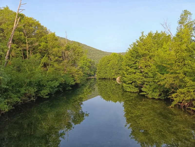 Photo d'un petit fleuve à l'eau calme, entouré d’arbres, avec des collines en arrière-plan et des reflets nets sur la surface. On distingue à peine le vestige d'un ancien moulin au centre-droit de la photo.