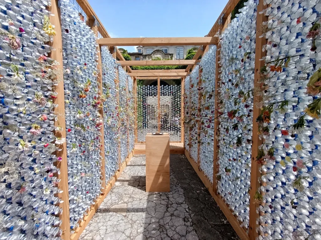 Photo d'un couloir extérieur formé par des murs en bois remplis de bouteilles en plastique transparentes et ornés de fleurs séchées, sous un ciel dégagé. Au centre, un petit meuble de bois avec un burger posé dans une vitrine.