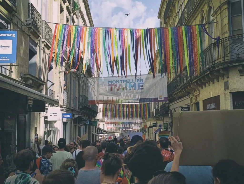 Photo d'une rue commerçante étroite à Montpellier remplie de personnes manifestant pour la Marche des fiertés, décorée de longues bandes de tissu arc-en-ciel suspendues et d’une banderole indiquant "Montpellier t’aime".