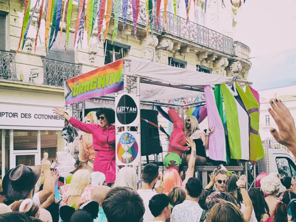 Photo d'un char de la marche des fiertés avec plusieurs drag queens en spectacle sur une estrade décorée de drapeaux LGBTQ+, entourée par une foule dans une rue commerçante de centre-ville.