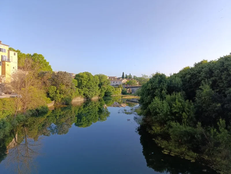Photo depuis le vieux pont d'un village gardois. Un petit fleuve bordé d’arbres, surtout sur la droite, avec un pont en pierre et des maisons en arrière-plan sous un ciel dégagé.