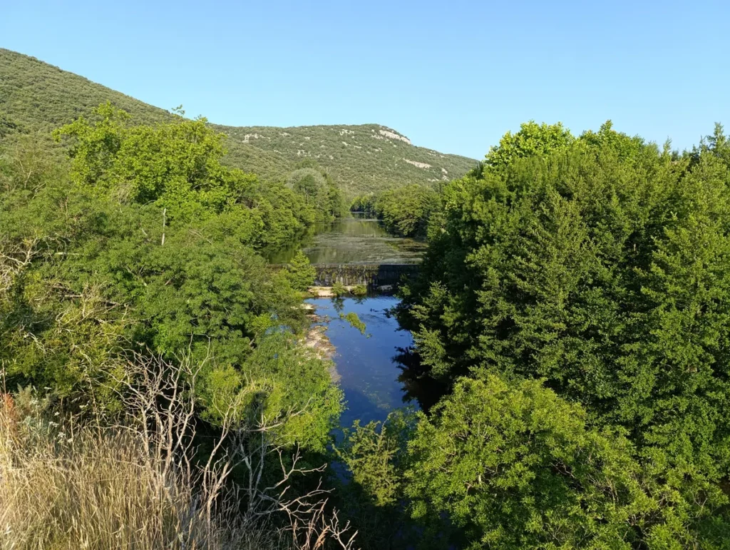 Petit fleuve traversant une vallée boisée, avec des collines en arrière-plan et des arbres denses de chaque côté. Une cascade artificielle entrave le cours d'eau au centre de l'image. Au dessus le ciel est bleu, sans nuage.
