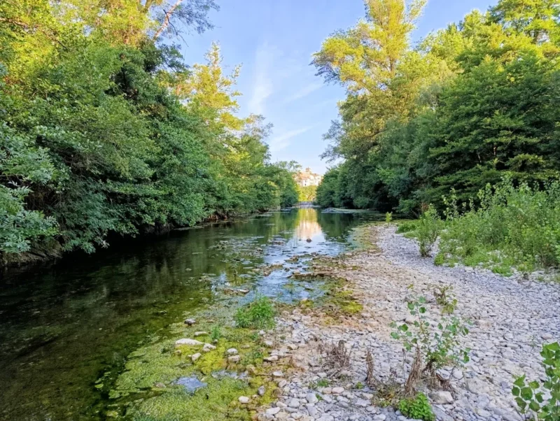 Photo d'un petit fleuve peu profond bordé de galets et de végétation, entouré d’arbres denses, avec les maisons d'un village visibles au loin au centre de l’image. Le ciel est dégagé.