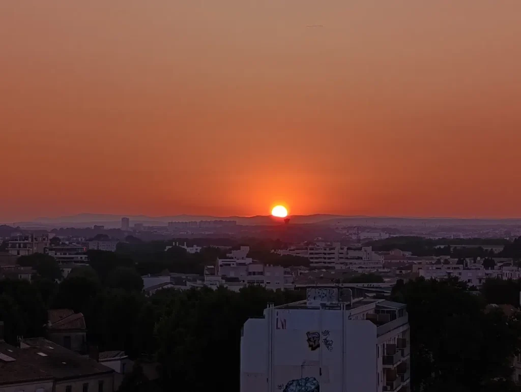 Photo d'une vue sur les toits de Montpellier, au coucher du soleil, sous un ciel dégagé. Le ciel est entre le jaune et le rouge, et on voit l'astre en train de disparaître au centre de l'image.