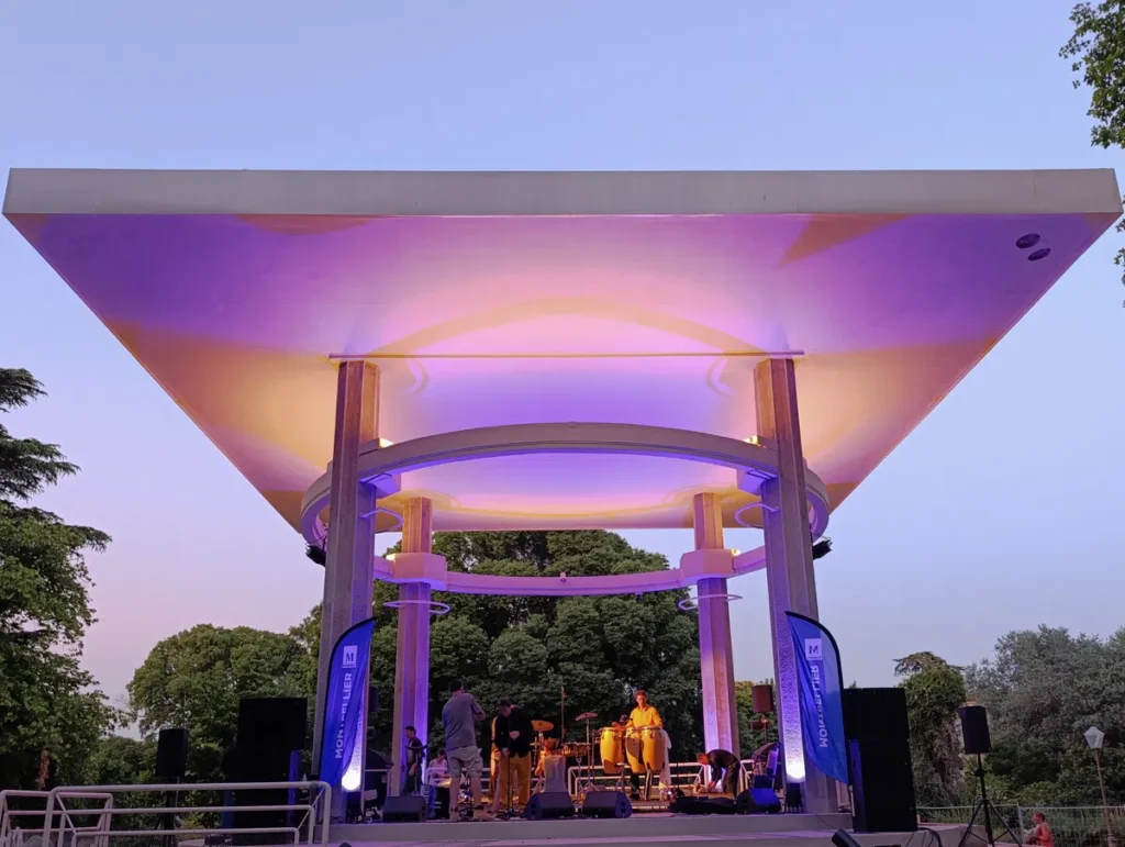 Photo d'un kiosque à musique moderne éclairé en violet, avec des musiciens sur scène au début d’un concert en plein air. Le ciel derrière est dégagé, la nuit tombante.