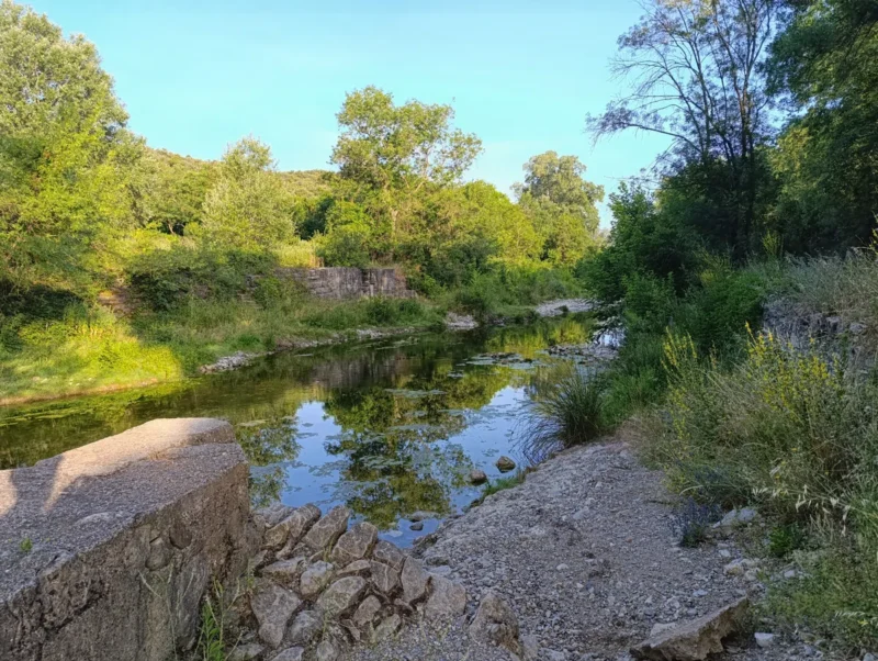 Photo d'un petit fleuve bordé de végétation dense, avec des ruines en pierre sur la rive gauche et des rochers en premier plan. Un mur vient se terminer sur le bord gauche de l'image. Au-dessus, le ciel est bleu, dans une lumière matinale.