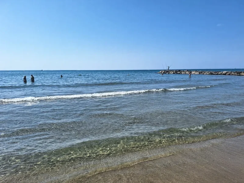 Photo d'une plage sous un ciel bleu. Quelques vaguelettes sur le sable au premier plan. Deux personnes debout, dans l'eau, sur la gauche de l'image. Sur la droite, on perçoit une digue de rochers.