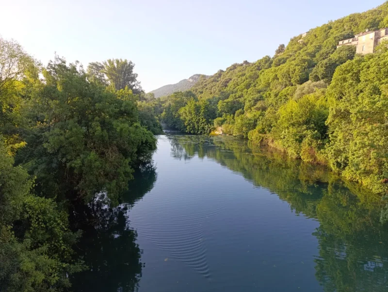 Photo prise d'un vieux pont, en aval d'un village. Vue d’un cours d'eau bordé d’arbres de chaque côté. Sur la droite, les dernières bâtisses du village. Le Ciel au-dessus est bleu.