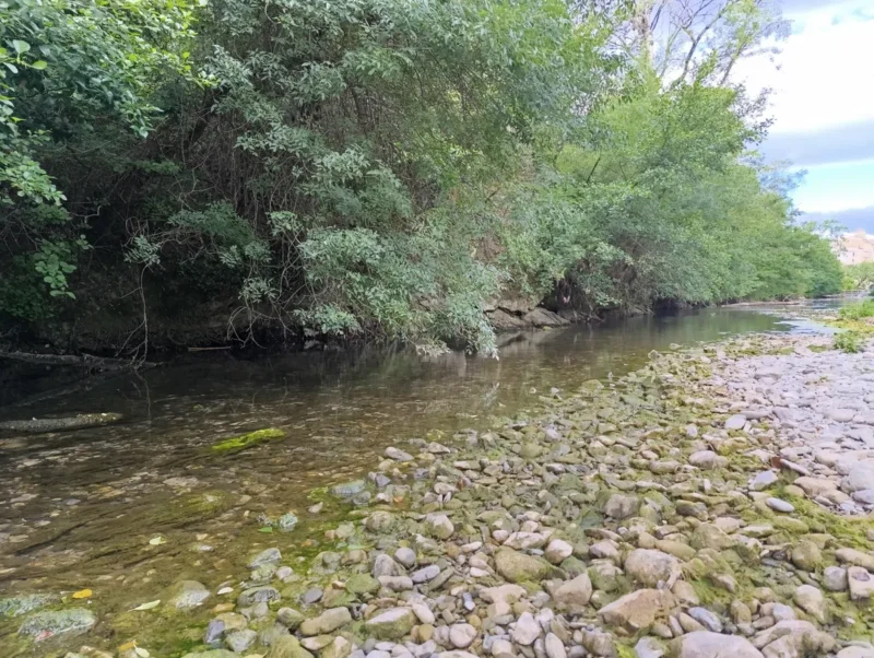 Photo d'un petit fleuve peu profond et bordé de galets. La rive opposée est densément bordée d’arbres aux feuillages épais. À l’arrière-plan, on distingue les dernières maisons d'un village. Le ciel est partiellement nuageux.