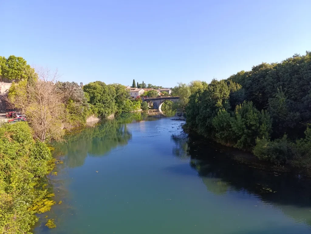 Un pont au loin sur un petit fleuve, le tout sous un ciel bleu. On perçoit quelques maisons d'un village et des voitures garées sur la gauche de l'image. Sur la droite, la végétation et les arbres sont à l'ombre d'une lumière matinale.
