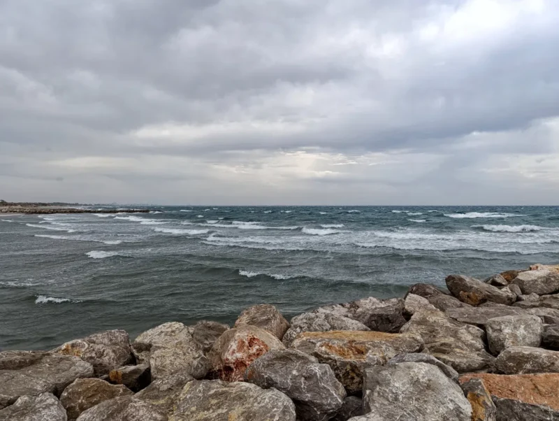 Photo d'une mer furieuse sous un ciel nuageux, derrière une digue de roches qui prolonge une plage non visible à l'image.