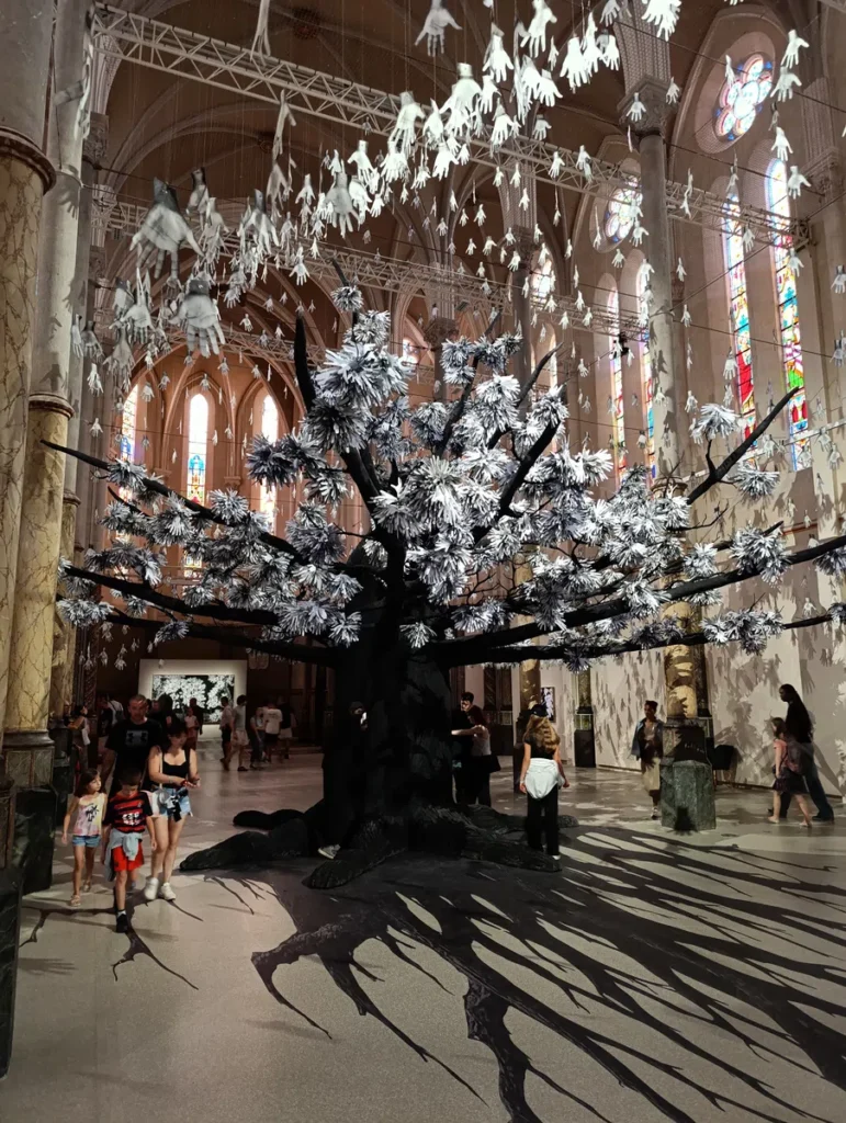 Photo prise dans l'église désacralisée Saint-Anne, à Montpellier. Posé au milieu de l'édifice, un gigantesque arbre dont les feuilles sont des mains de papier. Des visiteurs de l'exposition marchent tout autour. Au fond de l'église, on perçoit également une peinture.