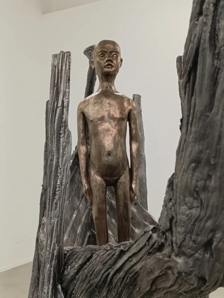Photo d'une sculpture couleur bronze représentant un enfant nu et debout, entouré de formes verticales semblant de bois, une barque.