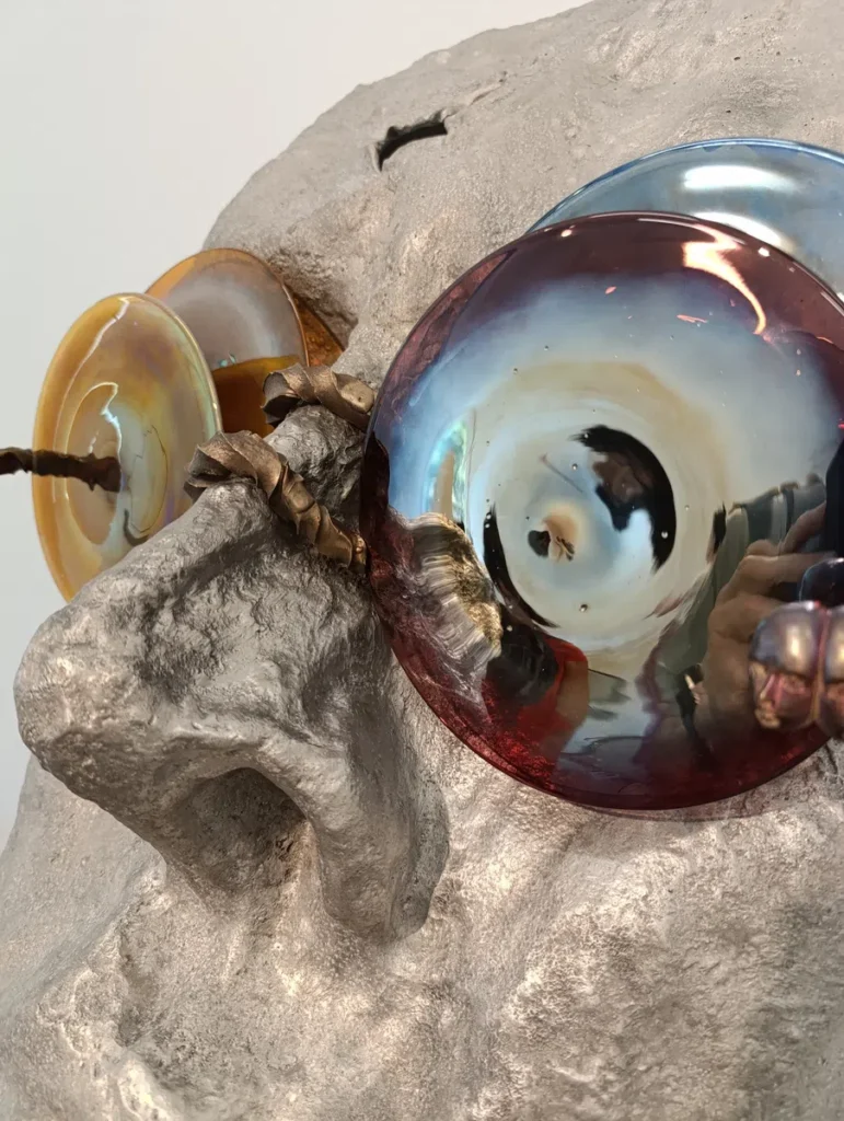 Photo en gros plan d’un visage sculpté en aluminium avec des disques en verre coloré fixés sur les yeux.