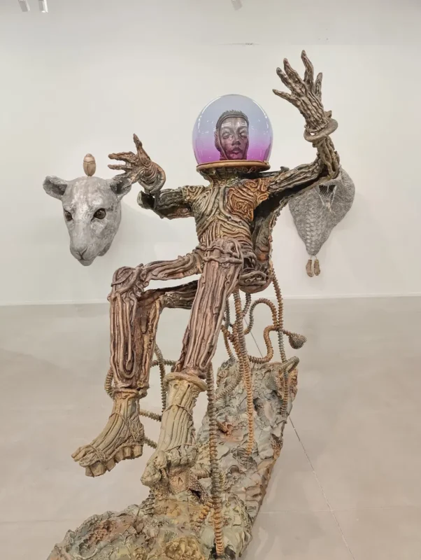 Photo d'une sculpture d’une figure humanoïde assise ou flottant en micro-gravité, au corps texturé comme du bois, avec une tête dans un casque sphérique transparent et des éléments décoratifs en arrière-plan, dont une tête de mouton.