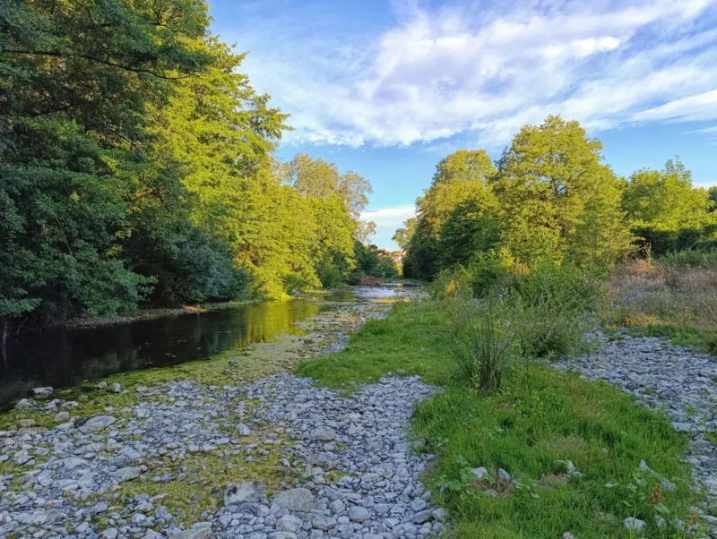 Photo d'un petit fleuve côtier bordé de végétation dense, avec sa plage de galets au premier plan. On peut distinguer au loin, sur ses rives, les premières maisons d'un village. Le ciel partiellement nuageux.