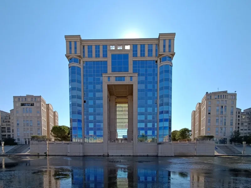 Photo d'un immeuble symétrique à façade vitrée bleue, encadré par deux bâtiments beiges, avec un plan d'eau au premier plan et un ciel dégagé en arrière-plan.
