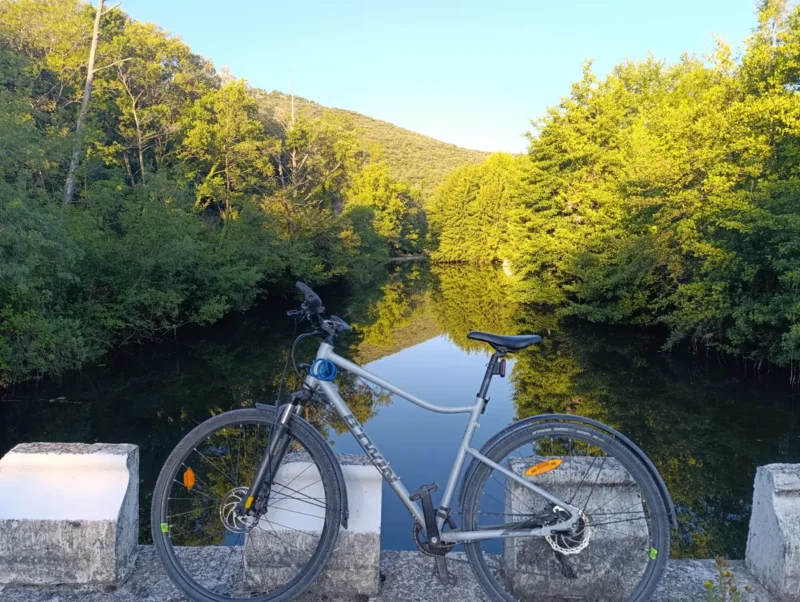 Photo d'un vélo gris au premier plan posé le long d'un muret en pierre sur un pont, avec en arrière-plan un petit fleuve calme entouré de végétation. Colline au loin et ciel bleu.