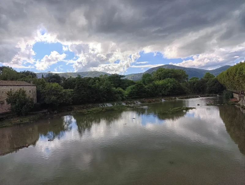 Photo d'un petit fleuve sombre et boueux sous un ciel nuageux. Quelques bouts de ciel bleu. Sur la gauche, une bâtisse. Sur la droite, on perçoit une rangée de platanes. Le cours d'eau est traversé par une petite cascade artificielle.